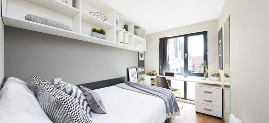 Light modern room with decor in tones of grey and white. Small double bed against the wall and opposite a window. White shelving on the wall above the bed, a desk and drawers under the window.