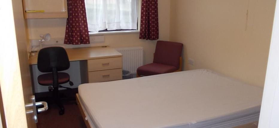 Double bed and desk with shelving. One window on the back wall with red curtains.