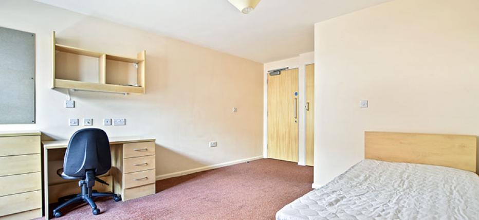 Large room with cream walls and red carpet, Single bed in the bottom left corner and desk, drawers and shelving along the right wall.