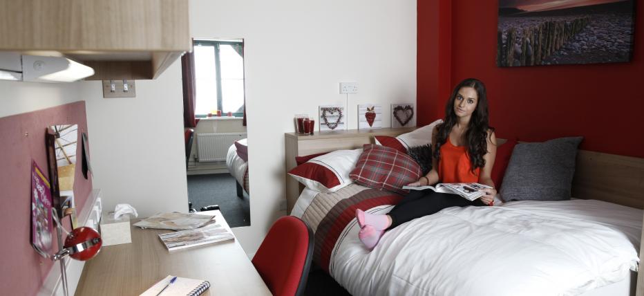 Student in a single room in Telford court, with a small double bed, desk, shelving, a noticeboard and a chair