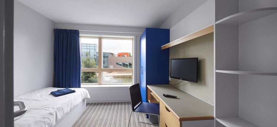 Bedroom interior showing single bed opposite a window overlooking campus. The room has a study area with wall mounted TV, drawers, wardrobe and shelves.