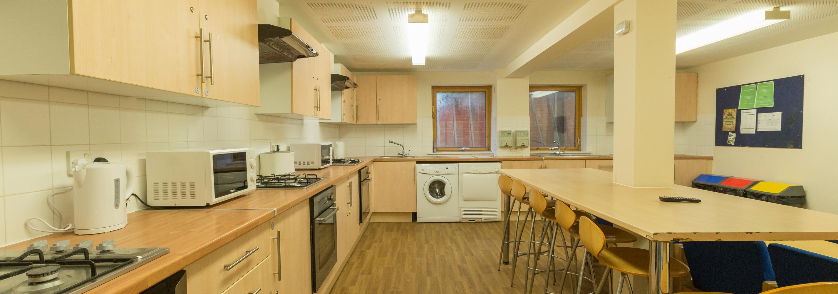 Kitchen, 3 gas cookers, 3 ovens, 3 extractor fans, 2 microwaves, 2 kettles, 2 windows, washing machine, dryer, noticeboard, toaster, dining table, 6 wooden bar stools, cupboards and bins