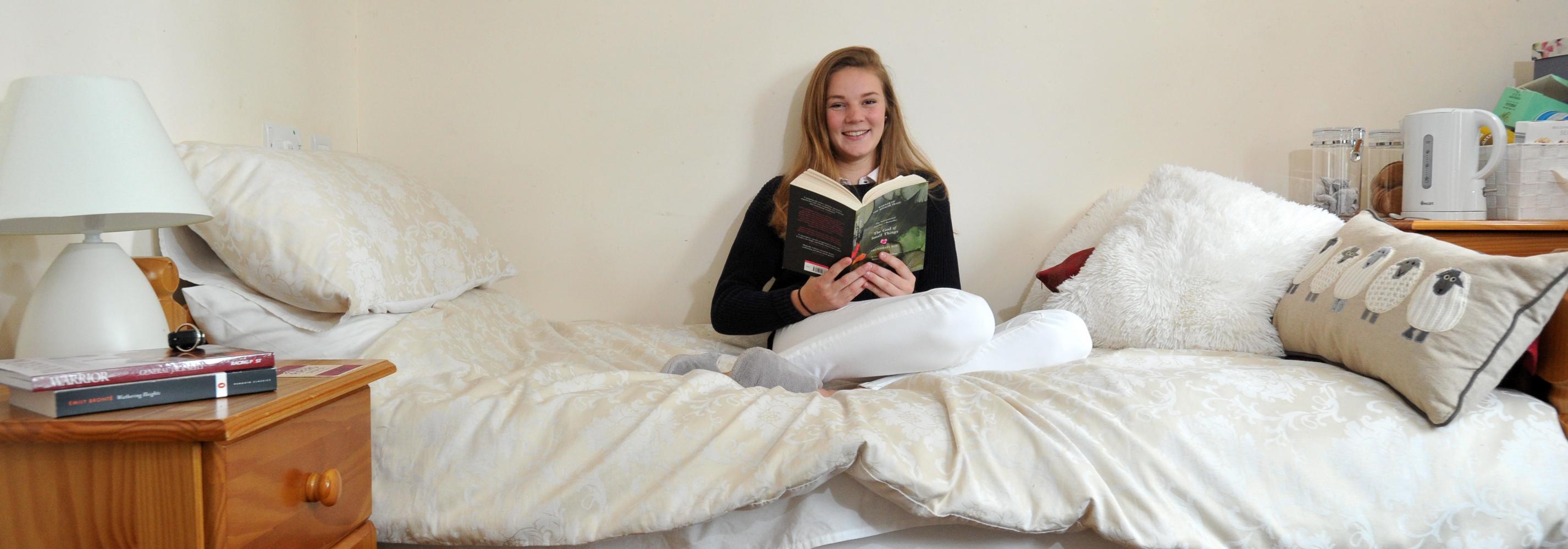 Student sat on bed reading. Room has a bedside table, storage and a noticeboard