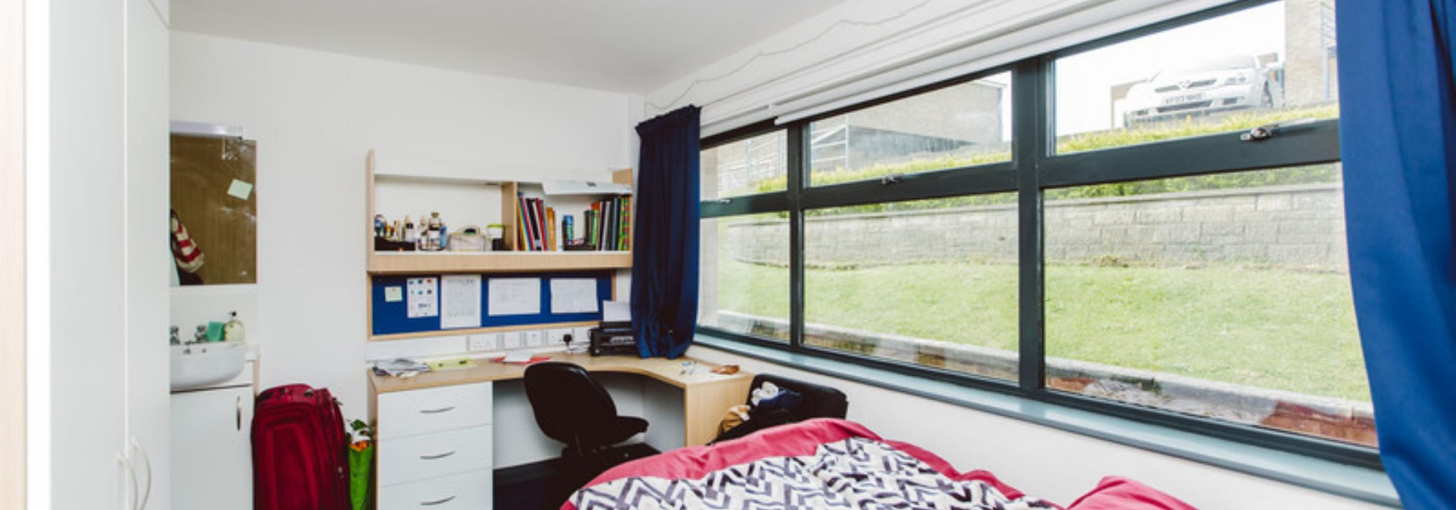 Large room with wide windows and blue curtains. A small double bed next to the window with the desk and shelving directly opposite.