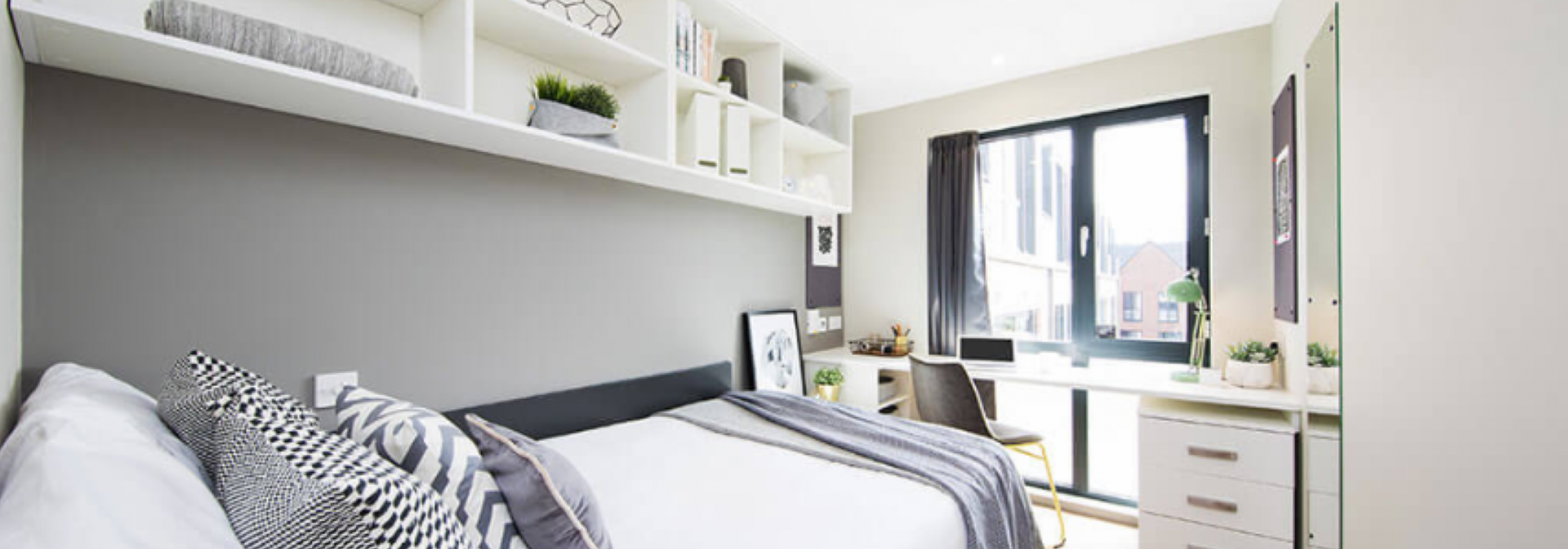 Light modern room with decor in tones of grey and white. Small double bed against the wall and opposite a window. White shelving on the wall above the bed, a desk and drawers under the window.