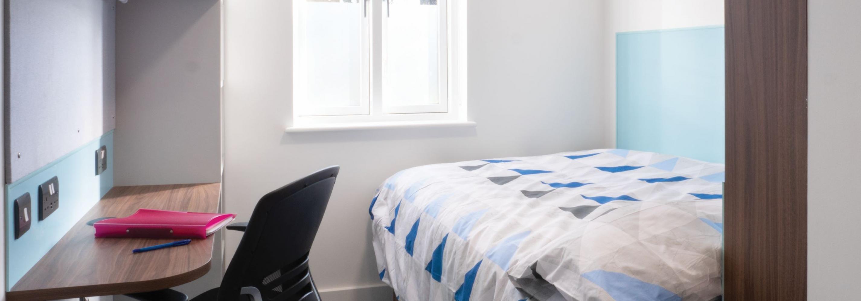 White bedroom with wood and blue accents. Small double bed facing the window. Blue blinds above the window. A desk, desk chair and wall shelving to the left.