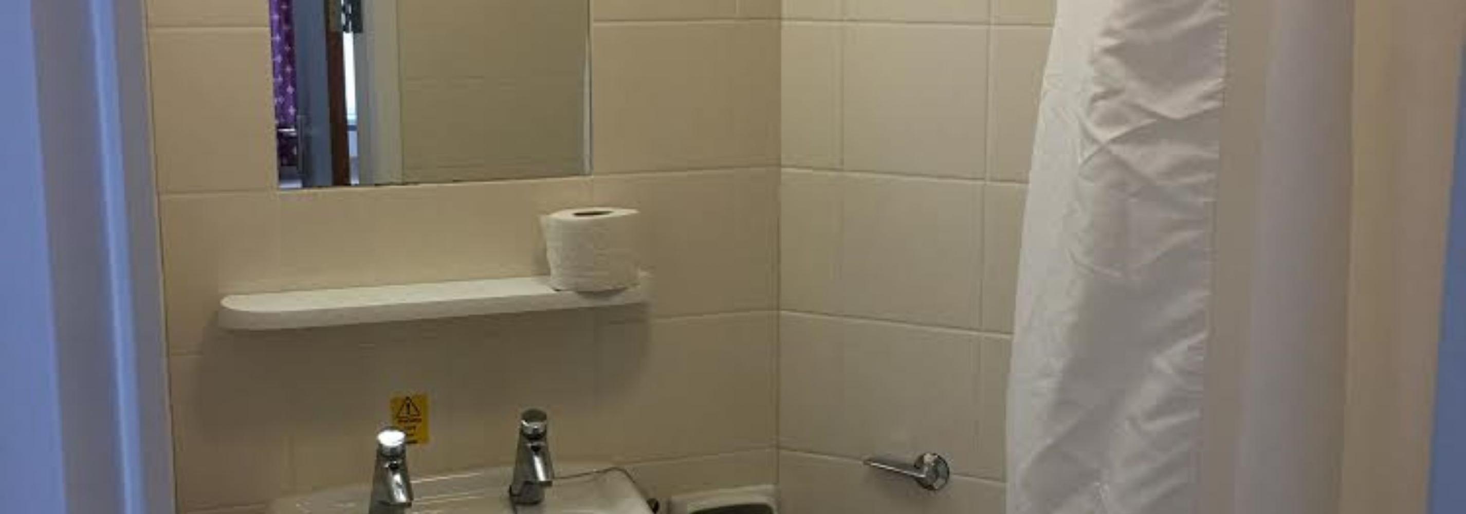White shower room with show curtain to the right, basin to the left with mirror and light above. Toilet in the middle.