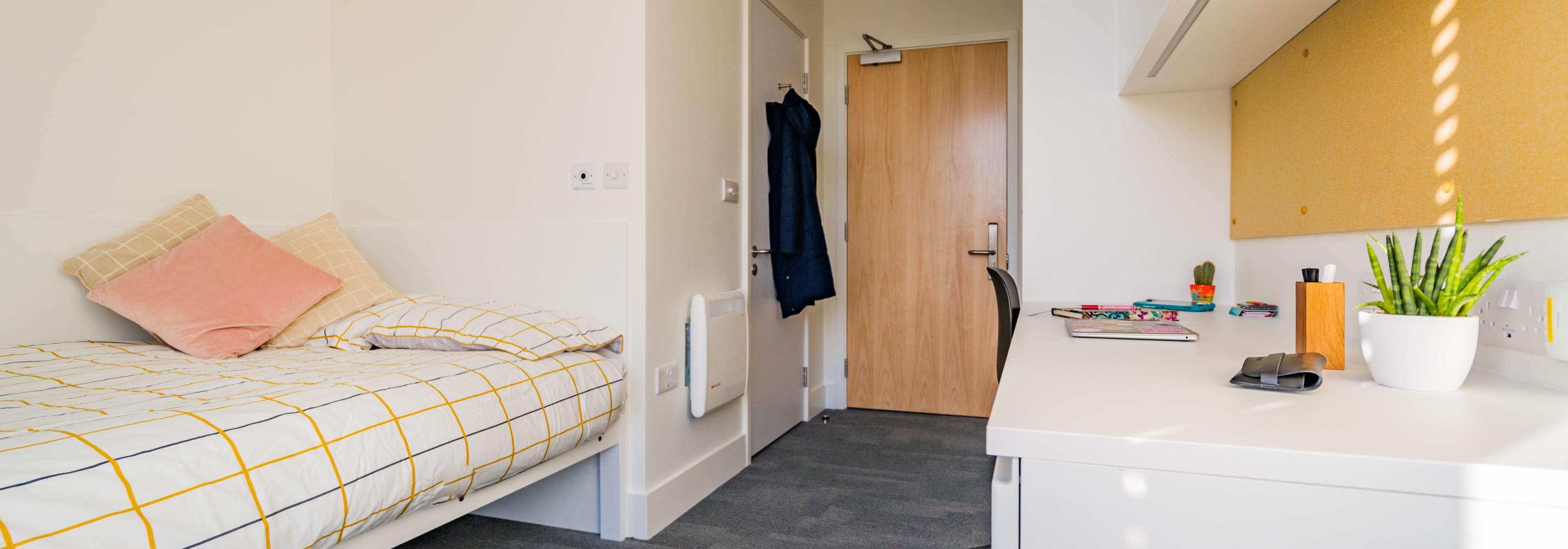A single en suite room showing a small double bed against one wall and a desk and wardrobe against the opposite wall. The en suite shower room is behind the bed with the door to the en suite next to the door into the room