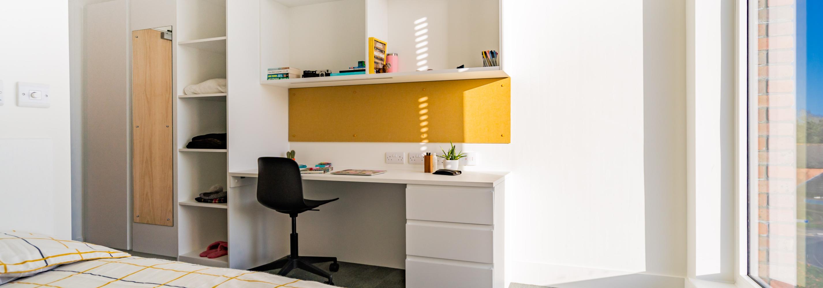 A typical single room with a large full height window and windowsill. A study desk and chair are next to the window with lots of storage space and a built in pinboard. Next to the desk is a floor to ceiling set of deep shelves and a wardrobe.