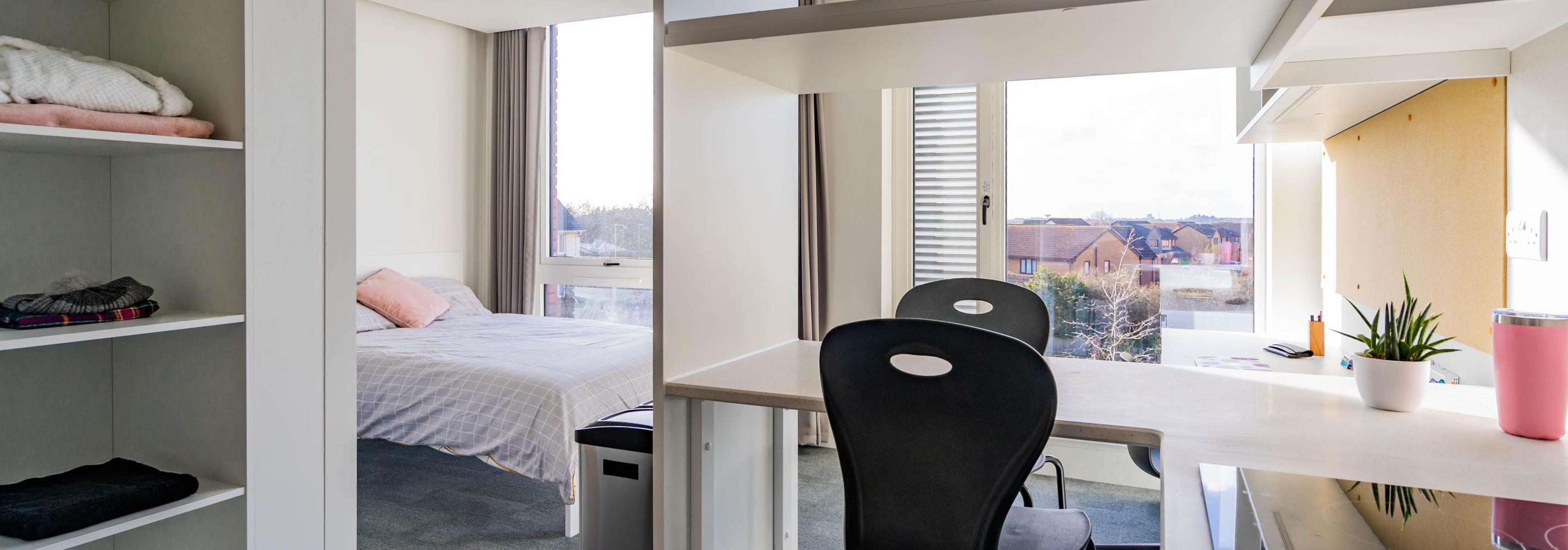 A self contained studio showing a double bed and study area next to floor to ceiling windows. A large set of storage shelves and a breakfast bar seating area with 2 seats is between the camera and the bed.