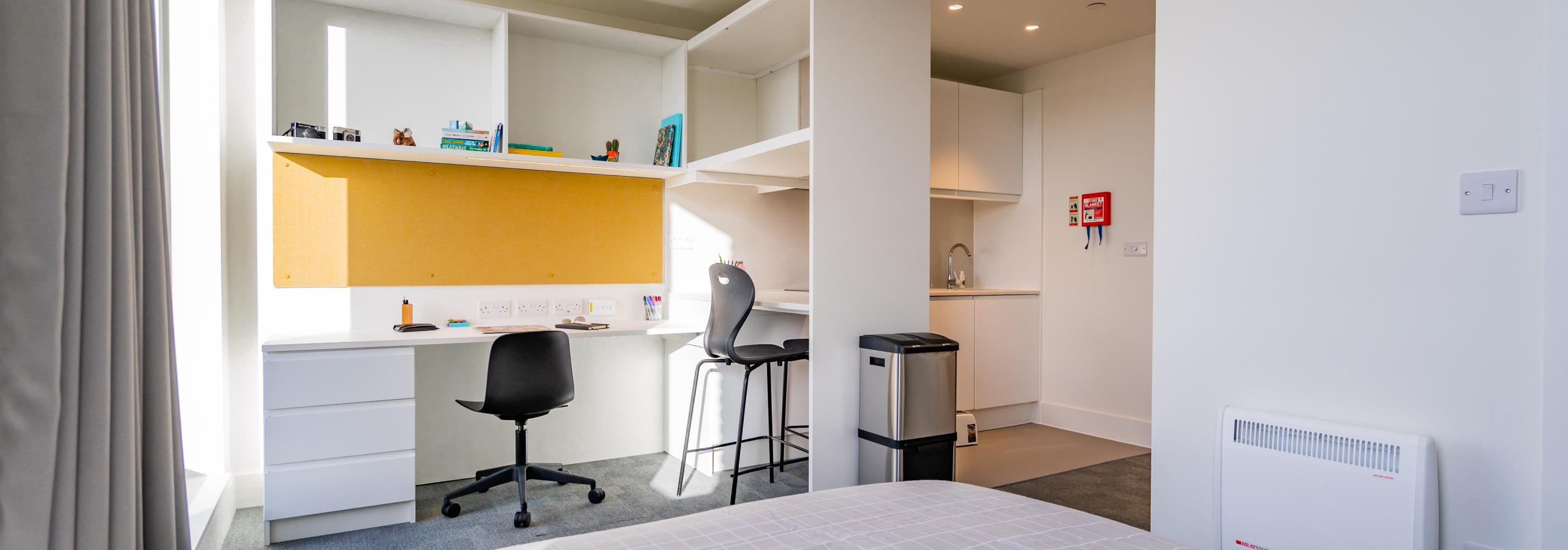 A self contained studio view from the double bed showing the large desk and study area with power and desk chair, the breakfast bar seating with storage shelves above and the small kitchen area.