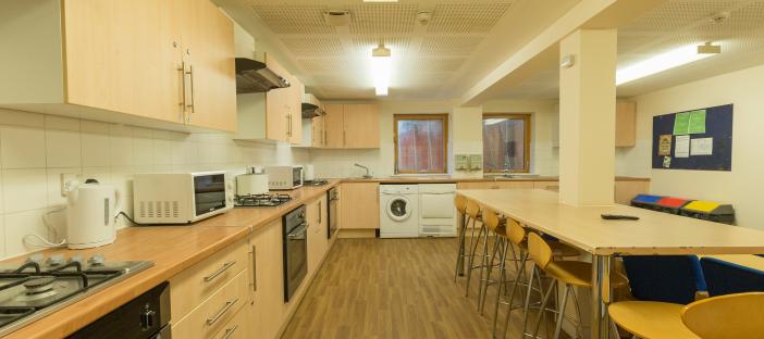 Kitchen, 3 gas cookers, 3 ovens, 3 extractor fans, 2 microwaves, 2 kettles, 2 windows, washing machine, dryer, noticeboard, toaster, dining table, 6 wooden bar stools, cupboards and bins