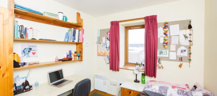 Room with cream walls. Single bed on the right of the room and desk, chair and shelving on the left. Window on the far wall with red curtains.