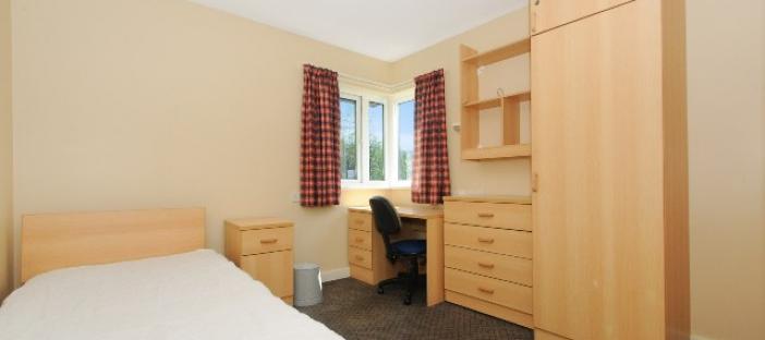Single bed on the left with a corner window and red curtains. Wooden wardrove, chest of drawers, study desk and shelving to the right. Wooden bed side table next to the bed.