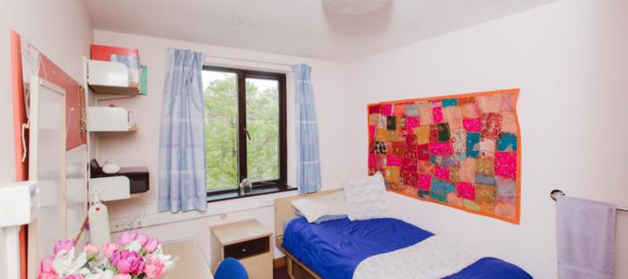 Single bedroom with bed to the right displayed with blue bedding and a pin board next to the bed. On the left there is a desk and shelving. On the far wall there is a window with light blue curtains.
