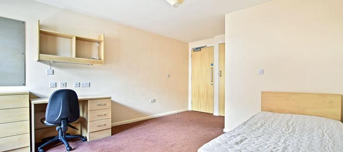 Large room with cream walls and red carpet, Single bed in the bottom left corner and desk, drawers and shelving along the right wall.