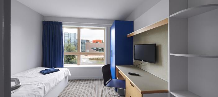 Bedroom interior showing single bed opposite a window overlooking campus. The room has a study area with wall mounted TV, drawers, wardrobe and shelves.