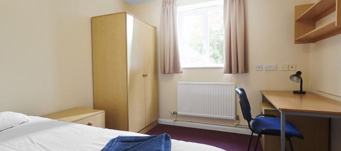 Bedroom interior showing single bed opposite a window with wardrobe, drawers, desk, chair and shelves. 