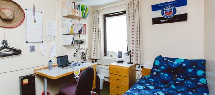 Bedroom with bed on the right side covered in a blue duvet, flag pinned above the bed, bedside table to the left of the bed and a study desk and chair on the left wall.